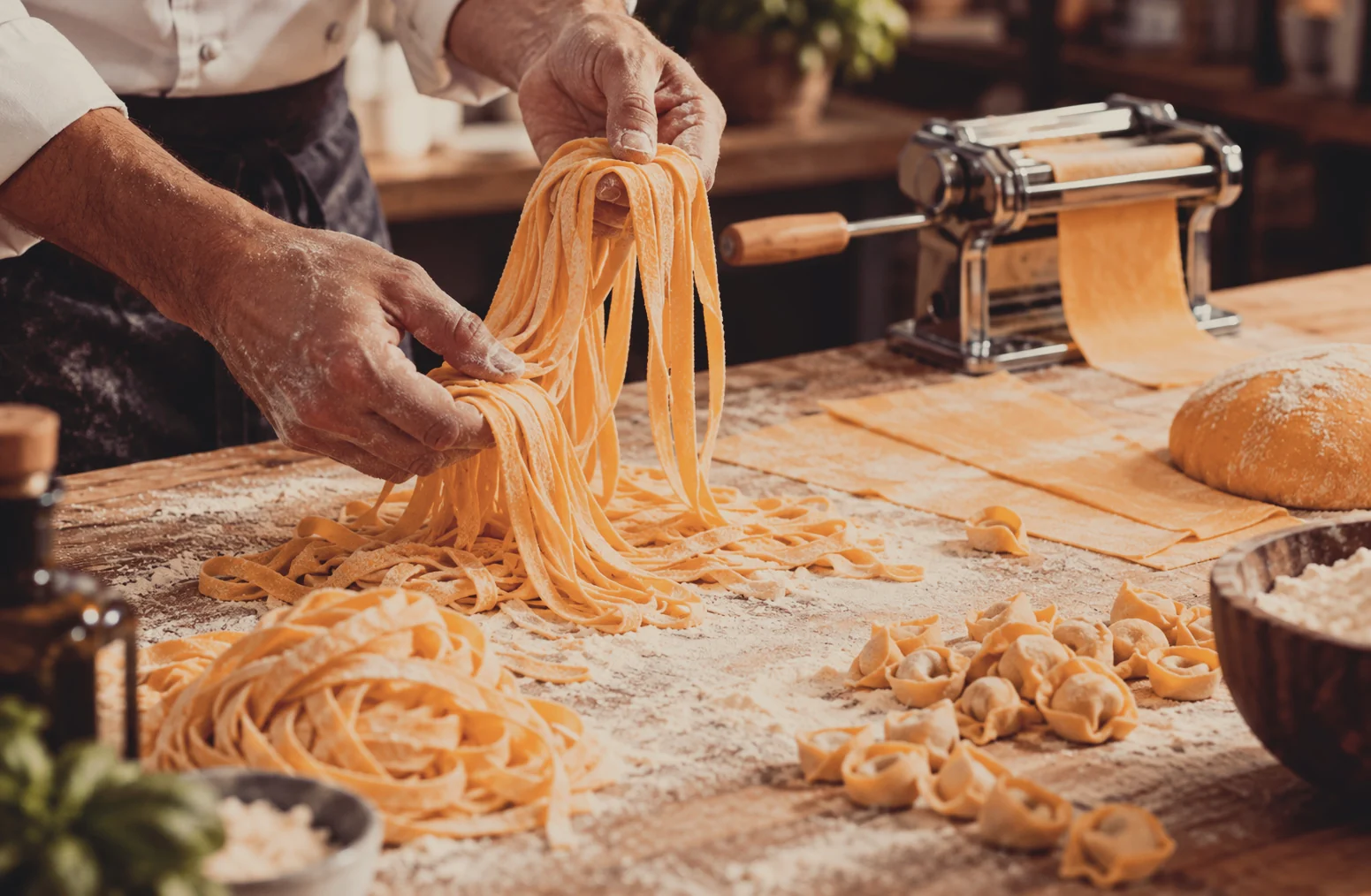 Frisch zubereitete Bandnudeln und Tortellini auf einem bemehlten Holztisch, dahinter eine Nudelmaschine